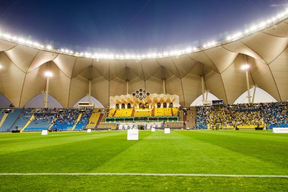 View from the pitch of King Fahd Sports City stadium in Riyadh, looking up towards people sitting in the stands and the tent-like walls opening to the evening sky